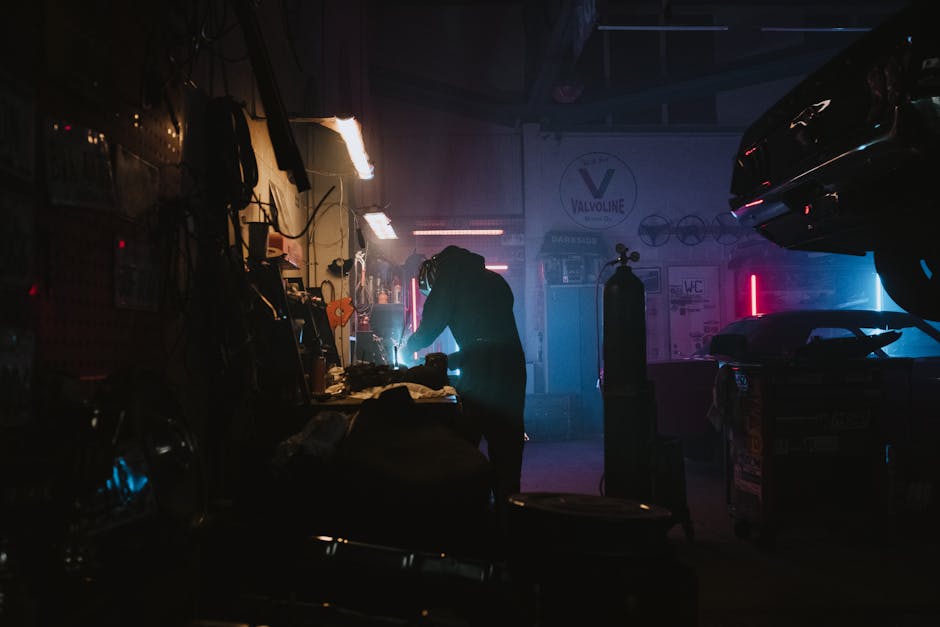 Silhouette of a mechanic working in a dimly lit garage, featuring neon lights and auto equipment.