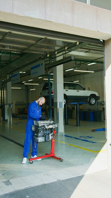 Mechanic in blue uniform inspecting an engine in a bright auto repair shop. Car lifted in background.