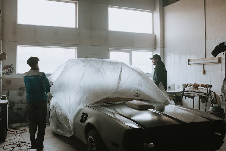 Two mechanics working on restoring a classic car in an auto garage with ample lighting.